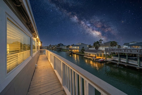A waterfront walkway with a view of the Milky Way, docked boats, and houses in the background.