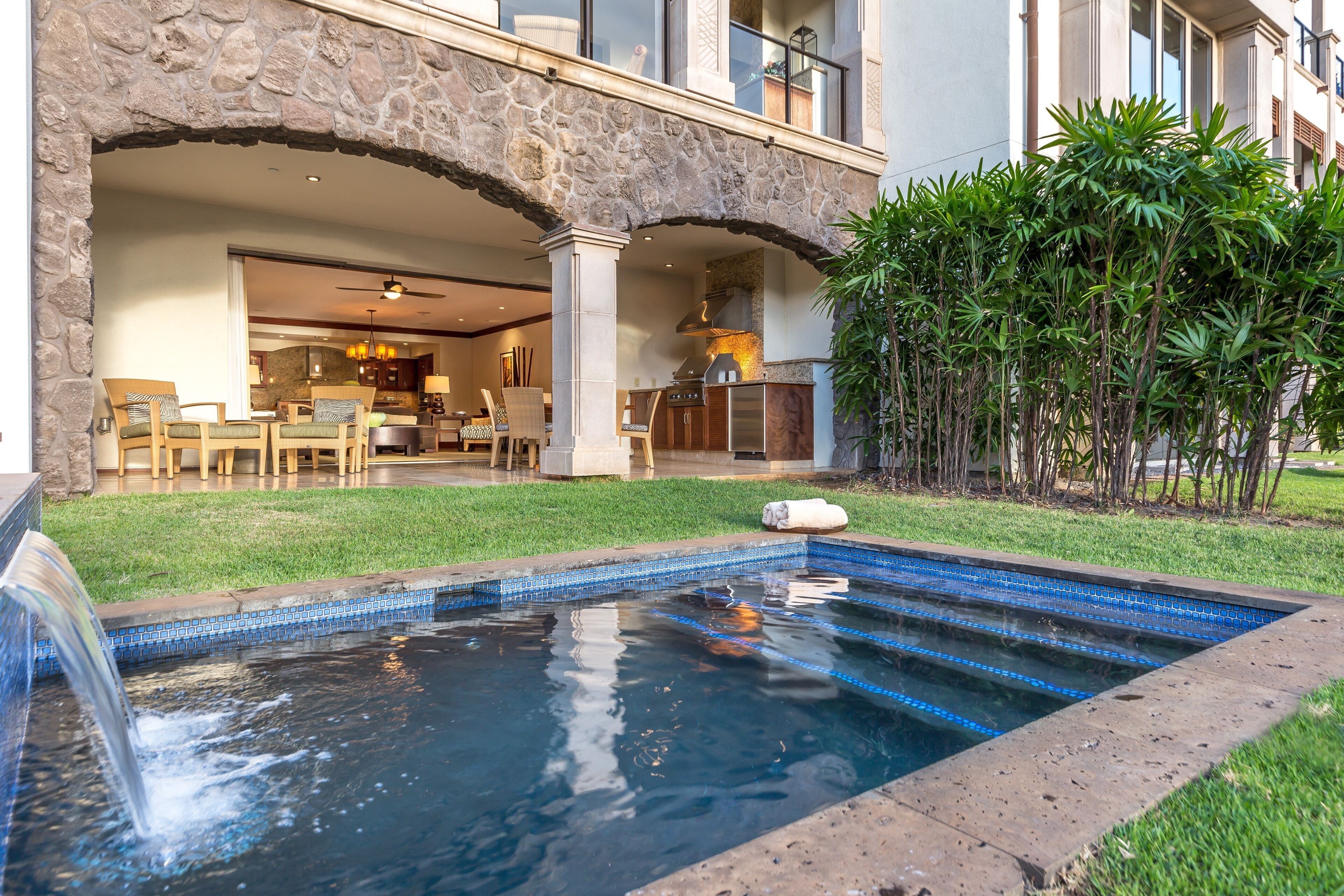 A swimming pool with a stone archway leading to a dining area, surrounded by lush greenery.