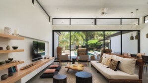Una sala de estar moderna con sofá, mesa de centro y sillas, un televisor y un ventanal con vista a una piscina y zonas verdes.