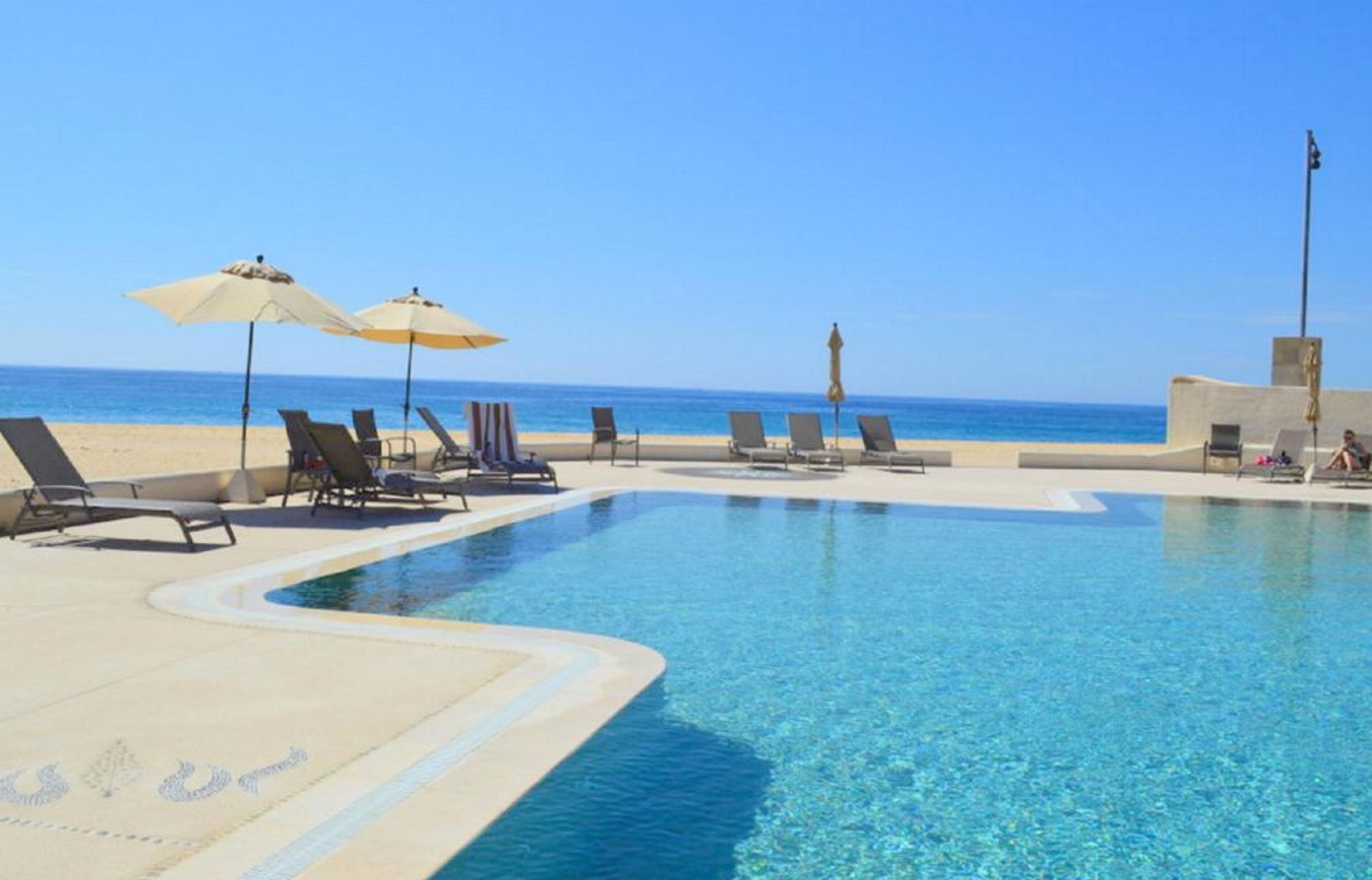 Una piscina azul cristalina con sillas de descanso y sombrillas, una playa arenosa y un cielo despejado.