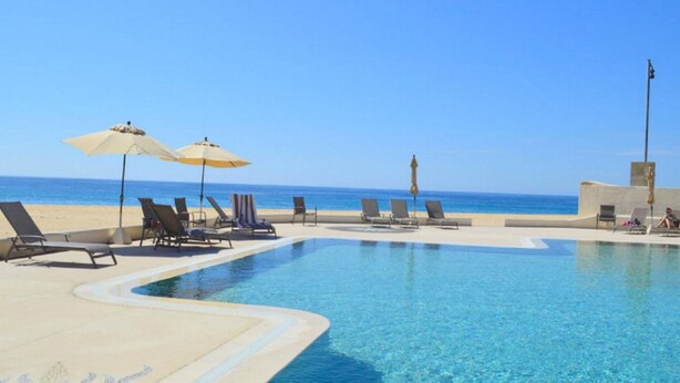 Piscina azul límpida com espreguiçadeiras e guarda-sóis, praia arenosa e céu claro.