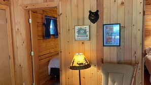 Interior de una cabaña de madera con una butaca acogedora, una mesa con lámpara y dos cuadros en la pared.