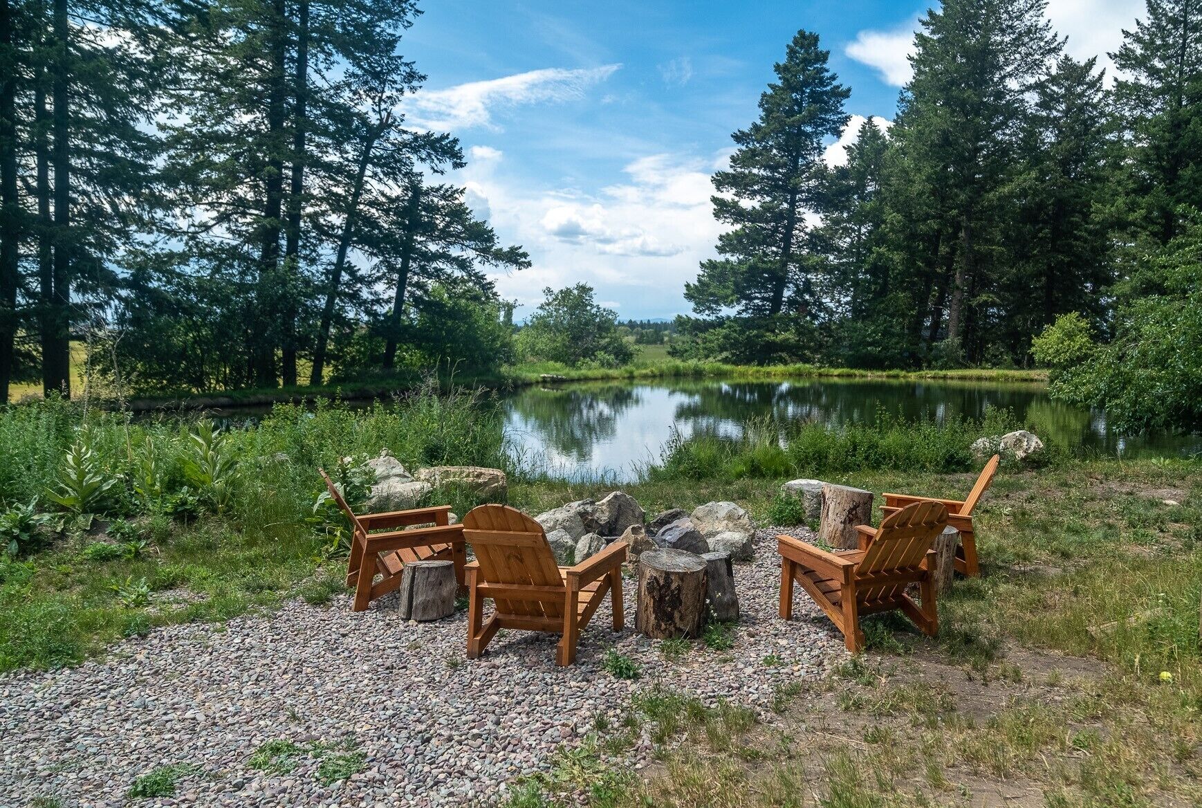 A lakeside seating area with wooden chairs and a fire pit surrounded by rocks.
