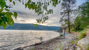 A rocky shoreline with a person standing near the water's edge, overlooking a calm lake with distant mountains. - Modern 3 bedroom, 3 en suites, dog friendly loft in McKinley (Kelowna)