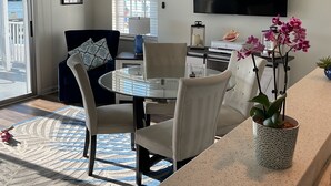 A modern living room with a glass dining table, white chairs, a blue sofa, a TV mounted on the wall, and a view of the outdoors through a sliding glass door. - Incredible Waterfront View, Pool, Dolphins, Fishing, Docks, Snook Inn 1 block up (Marco Island)