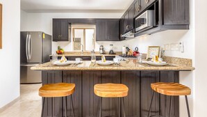 A modern kitchen with dark cabinetry, a stainless steel refrigerator, a microwave, a gas stove, and a granite countertop. There are three wooden stools at the bar area. - Casa Rosas - Private pool (Cabo San Lucas)