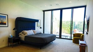 A modern bedroom with a large bed, a nightstand, a chair, and a view of the outdoors through large windows. - Quiet and peaceful retreat (La Quinta)