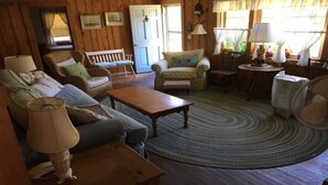 A cozy living room with a sofa, armchair, and coffee table. There is a door, a window with curtains, and a fan on the floor. - Old Cape Cod Charm in Chatham MA, 1/4 mile Walking Distance to Hardings Beach (Chatham)