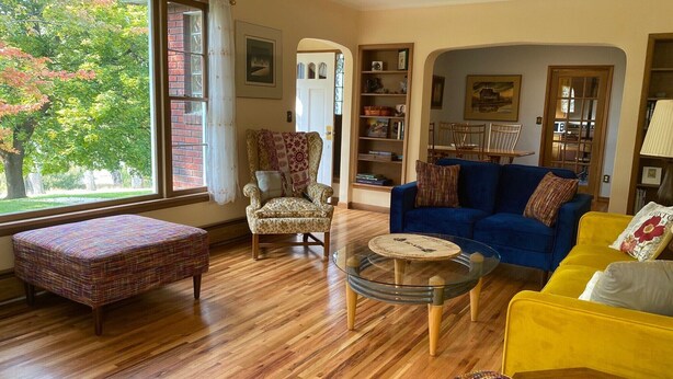 Een woonkamer met een groot raam, een blauwe bank, een gele fauteuil, een gestreept fauteuil, een glazen salontafel en een houten vloer.