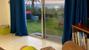Una sala de estar con sofá, una mesa de centro y una estantería. Hay un ventanal corredero que da a un patio con césped y una palmera.