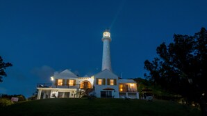 Un faro con una torre blanca y una luz encendida en la parte superior, rodeado por una casa con múltiples ventanas y un balcón.