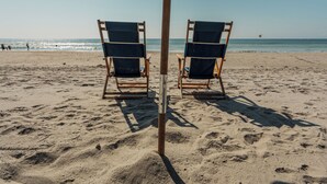 Une plage avec deux chaises de plage en bois sous un grand parasol sombre, une plage de sable et l’océan en arrière-plan.