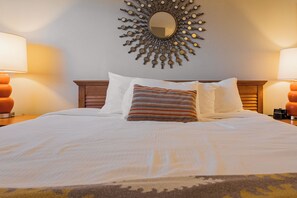 A neatly made bed with white linens, a patterned throw pillow, and a sunburst mirror above it. - North Myrtle Beach | Beach Escape | Pool & Ocean Access | 1BR Villa (North Myrtle Beach)