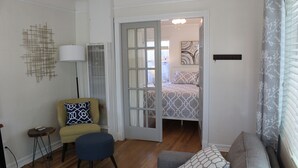 A modern living room with a sofa, armchair, and ottoman. A door leads to a bedroom with a patterned bedspread. - Charming North Park house, with parking and new central Air.  (San Diego)