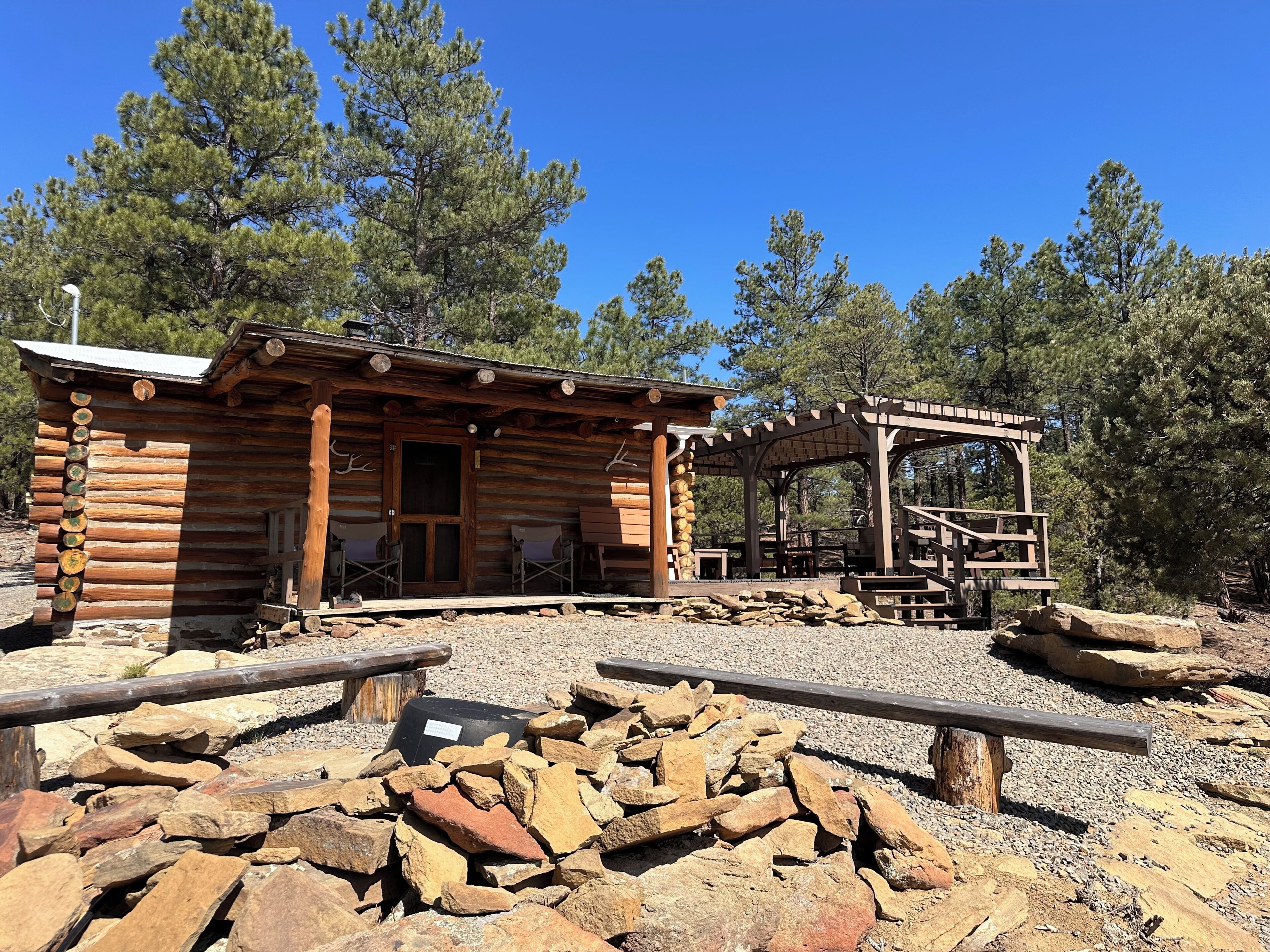 Une cabane rustique en rondins de bois, dotée d’une véranda, de sièges extérieurs et d’un espace abrité avec un escalier.