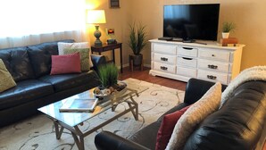 A living room with a black leather sofa, a glass coffee table, a white dresser, a flat-screen TV, and a potted plant.