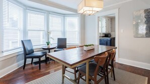 A dining area with a wooden table and chairs, a hanging light fixture, and large windows with blinds. - Charming Hideaway in Westport 15 (KCMO)