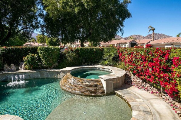 A hot tub surrounded by a landscaped garden with a pool, trees, and a mountain backdrop.