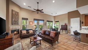 A spacious living room with a fireplace, a ceiling fan, and a dining area with a view. - Villa Puerta Azul · Backyard Oasis at Puerta Azul (La Quinta)
