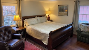 A bedroom with a large bed, a wooden headboard, a nightstand with a lamp, a leather armchair, and a window with curtains.