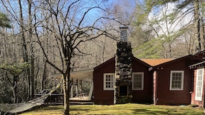A red wooden cabin with a stone chimney, surrounded by a grassy yard and trees.
