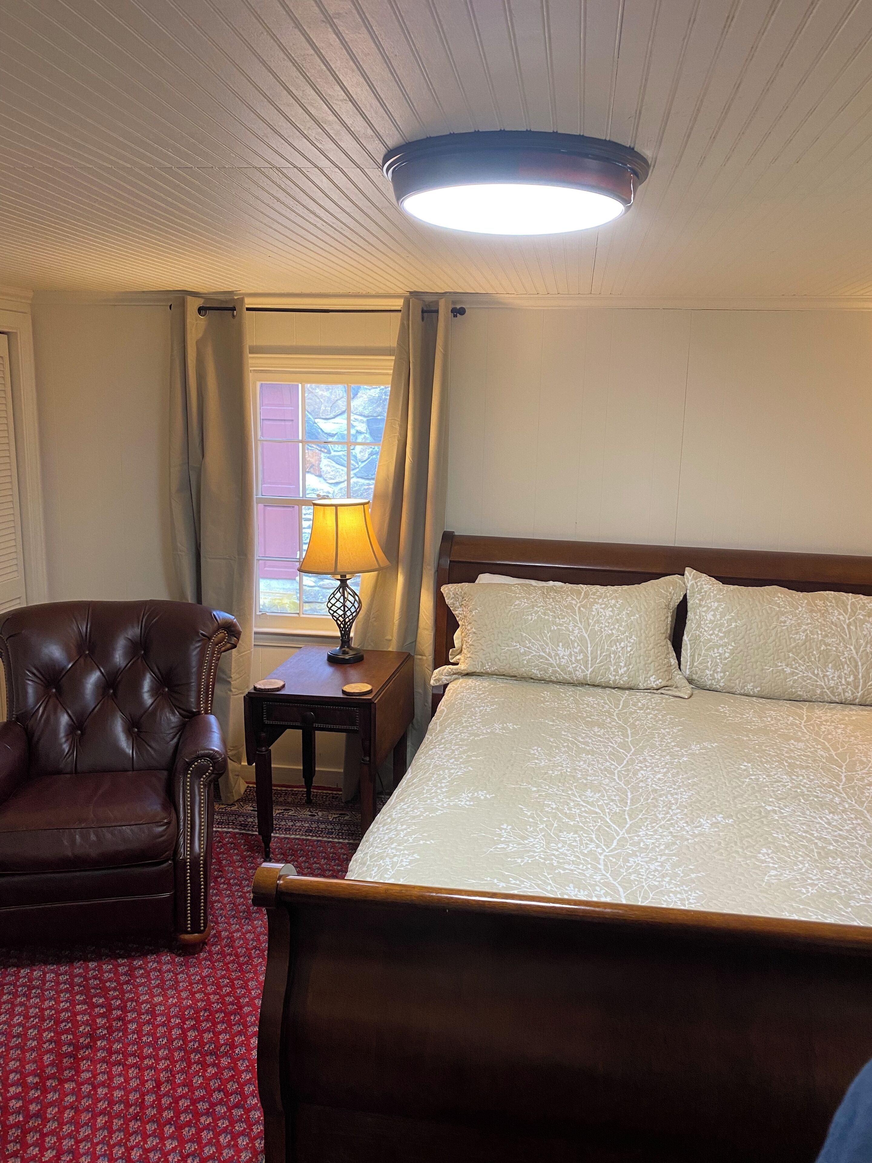 A hotel room with a large bed, a leather armchair, a nightstand with a lamp, and a window with curtains.