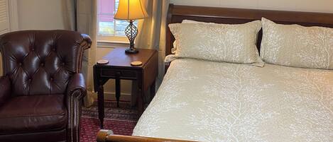 A hotel room with a large bed, a leather armchair, a nightstand with a lamp, and a window with curtains.