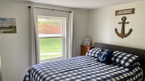 A bedroom with a checkered bedspread, a view of a lake through a window, a wooden nightstand, and a wall-mounted anchor decoration. - Cozy family & pet friendly lake home, great reviews & rates (Holcombe)