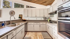 Een moderne keuken met witte kastjes, roestvrijstalen apparatuur, een houten plafond en een groot raam met uitzicht op bomen.