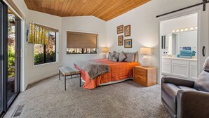 Een slaapkamer met een bed, nachtkastjes, een bankje, een stoel, een groot raam, een plafond met houten balken, en een badkamer met een spiegel en verlichting.