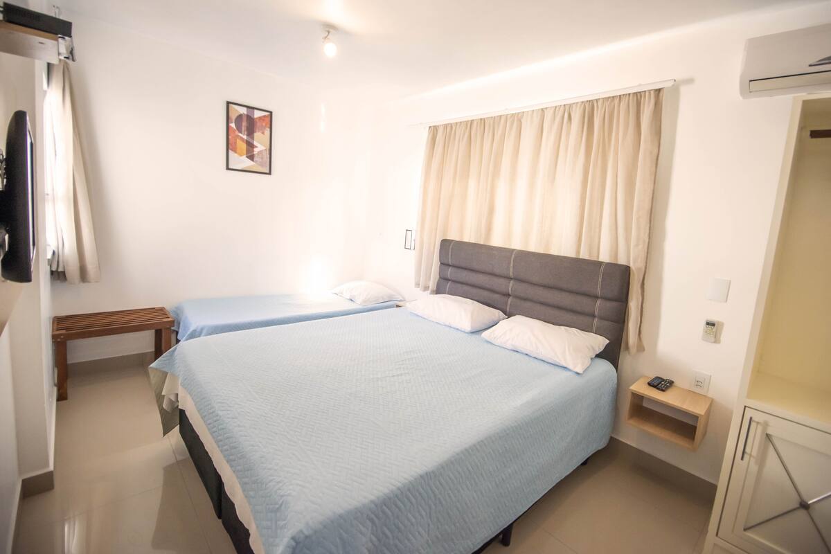 A hotel room with two beds, a bedside table with a phone, a small shelf, and a wall-mounted air conditioner.