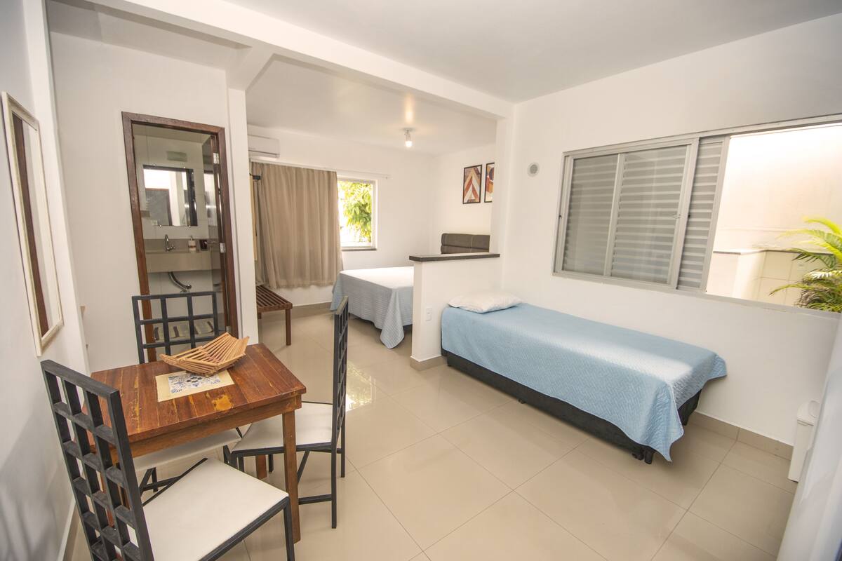 A compact room with a wooden dining table, a single bed, a small sofa, and a window with blinds.