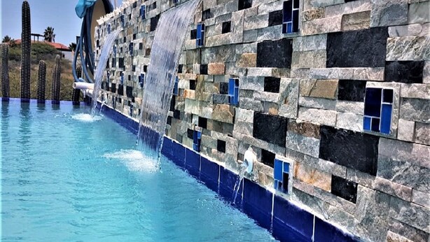 A pool with a waterfall feature and a stone wall with blue-tinted windows. - Superior 1-Bedroom Apartment - Aruba Cunucu Residence (Oranjestad)