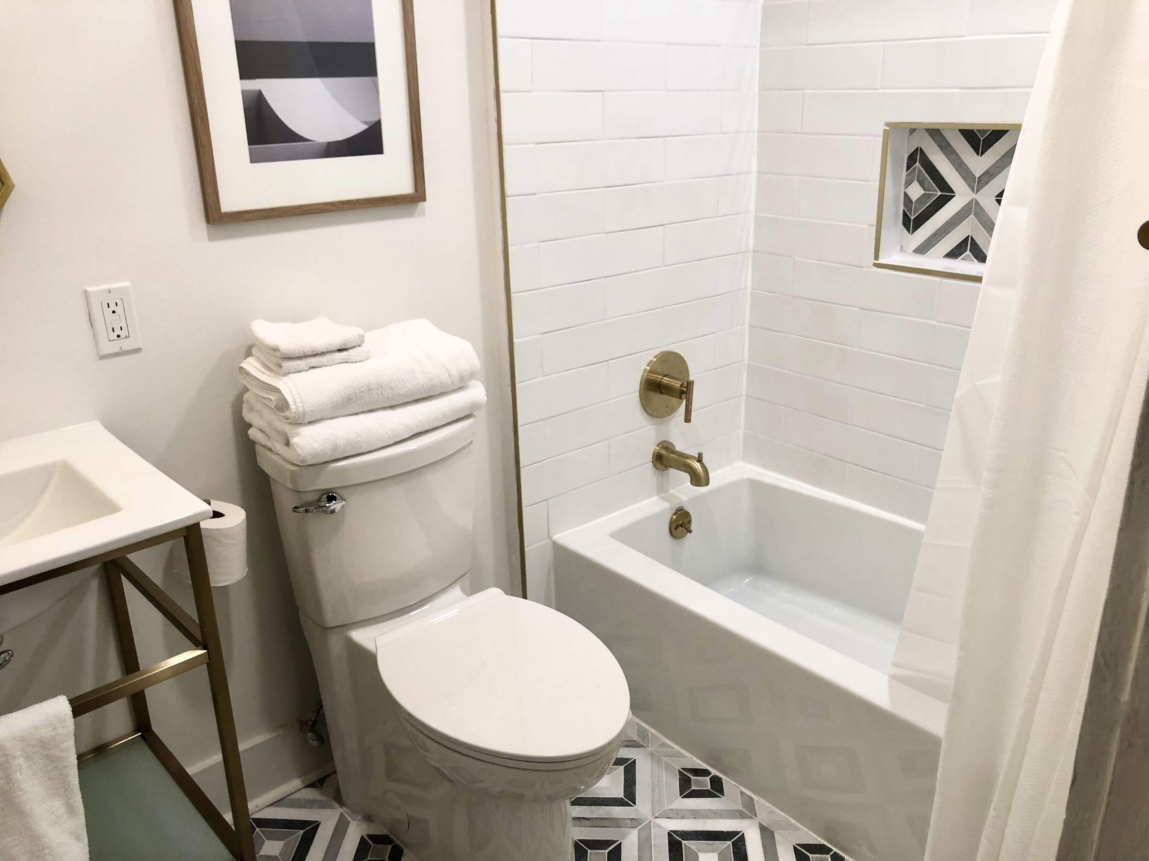 Une salle de bain avec des toilettes, un lavabo et une baignoire. Un tableau encadré est accroché au mur, et le sol est recouvert de carreaux disposés selon un motif géométrique.