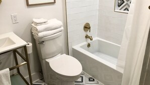 Un baño con inodoro, lavamanos y bañera. Hay un cuadro en la pared, y el suelo tiene un diseño de baldosas geométricas.