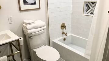 Une salle de bain avec des toilettes, un lavabo et une baignoire. Un tableau encadré est accroché au mur, et le sol est recouvert de carreaux disposés selon un motif géométrique.