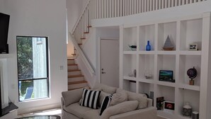 Een ruime woonkamer met een beigekleurige zithoek, een glazen salontafel en een trap met een witte leuning naar een hoger niveau.