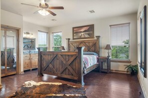 A bedroom with a wooden bed, a ceiling fan, a large window with blinds, a painting, and a rustic rug. - Lake View Retreat at CrossTimbers Marina (Sperry)