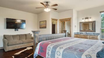 Un dormitorio con cama, ventilador de techo, televisión, sofá, cómoda de madera y baño con espejo y lavamanos.