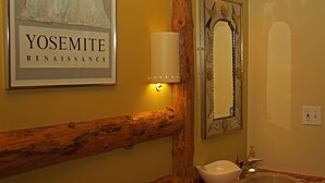 Ein Badezimmer mit Geländer aus Holzstämmen, einem gerahmten Kunstwerk mit dem Titel 'YOSEMITE RENAISSANCE', einer Wandleuchte, einem dekorativen Spiegel und einem Waschbecken mit Armatur.