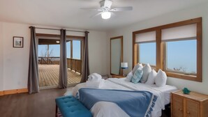 Ein Schlafzimmer mit einem großen Bett, einem Deckenventilator, einer hölzernen Sitzbank und Blick auf die Terrasse und den Himmel durch die Fenster.