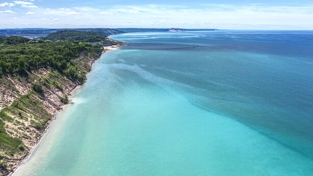 Luftaufnahme einer Küstenregion mit klarem türkisblauem Wasser, einem Sandstrand und einer bewaldeten Klippe.