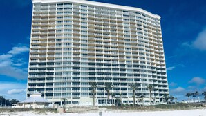 Een groot, meerhoog hotel met een gebogen gevel, gelegen aan een strand met palmbomen en een helderblauwe lucht.