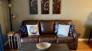 Ein Wohnzimmer mit einer braunen Ledersofa, zwei Zierkissen, einem gewebten Korb auf einem dunklen Couchtisch und drei Wandbildern über der Couch.
