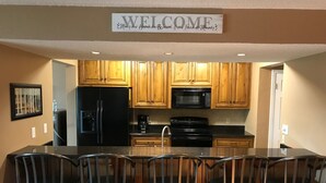 Eine Küche mit Holzausstattung, einem schwarzen Kühlschrank, einer Mikrowelle und einem Herd. Über den Schränken hängt ein Schild mit der Aufschrift 'WELCOME'.