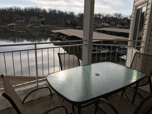 A balcony with a glass table and chairs, overlooking a body of water and buildings.