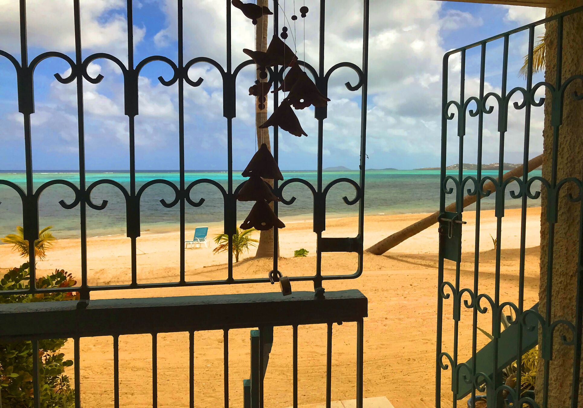 A beach view through a metal gate with decorative elements, a palm tree, and a chair on the sand.