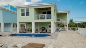 Ein zweistöckiges Haus mit Swimmingpool, einer überdachten Terrasse mit Liegestühlen und einer Holzterrasse.