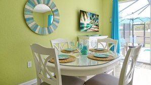 Een eetgedeelte met een ronde glazen tafel, witte stoelen en een tropisch schilderij aan de muur.
