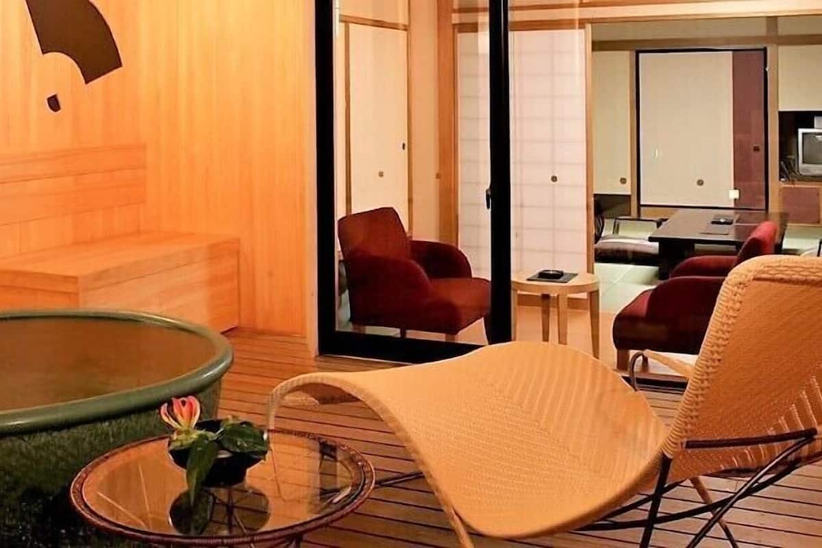 A room with a wooden floor, a large round table with a glass top, a red chair, a beige chair, and a small table with a flower vase.